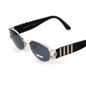 May include: Oval-shaped sunglasses with a silver frame and dark lenses. The temples are black with silver stripes and a circular logo. The sunglasses have a classic, retro design.