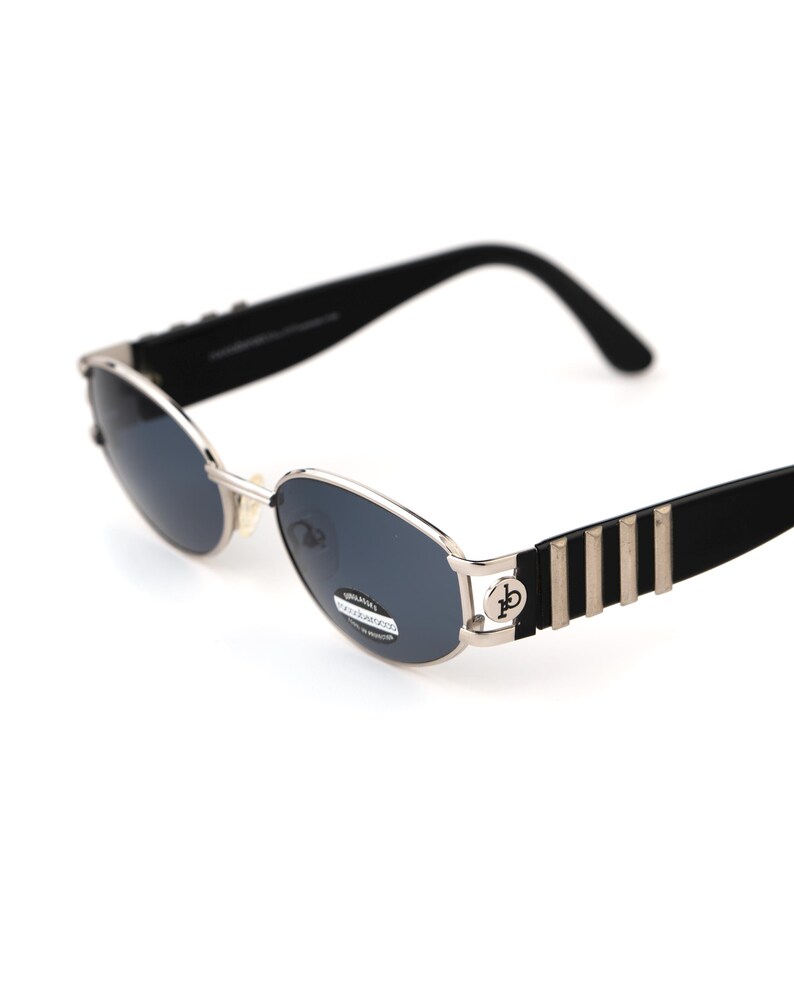 May include: Oval-shaped sunglasses with a silver frame and dark lenses. The temples are black with silver stripes and a circular logo. The sunglasses have a classic, retro design.
