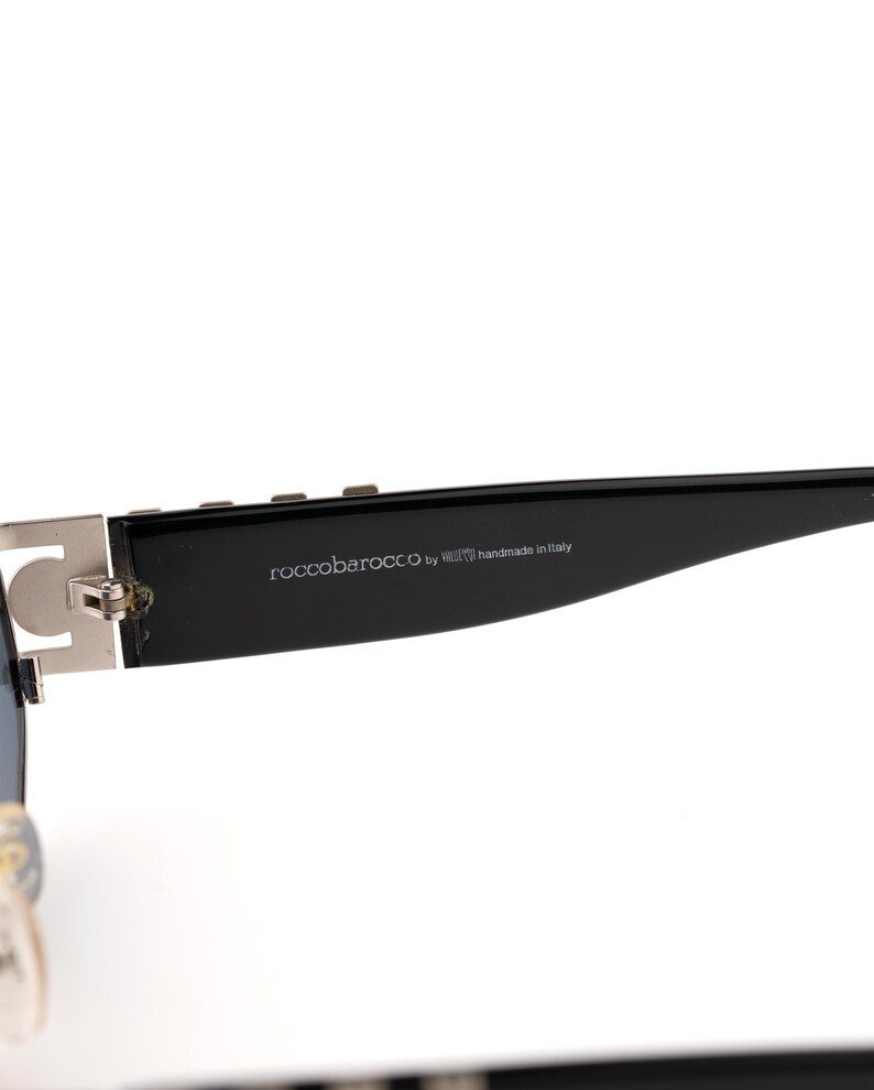 May include: Close-up of a pair of sunglasses. The sunglasses have a black frame with silver accents and the text "roccobarocco by handmade in Italy" printed on the arm.