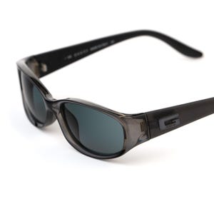 Puede incluir: Gafas de sol grises oscuras con montura rectangular y lentes oscuras. Las gafas tienen una montura sutil y translúcida y un pequeño logotipo en la patilla. El diseño es elegante y moderno.