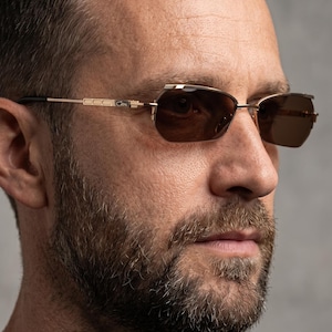 May include: Gold-framed sunglasses with brown tinted lenses. The sunglasses have a rectangular shape and a bridge over the nose. The temples are thin and extend to the ears. The sunglasses are worn by a person with a beard.