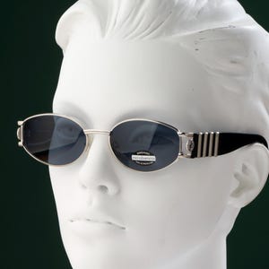 May include: Oval-shaped sunglasses with silver frames and dark lenses. The temples are black with silver stripes. The sunglasses are displayed on a white mannequin head against a dark green background.
