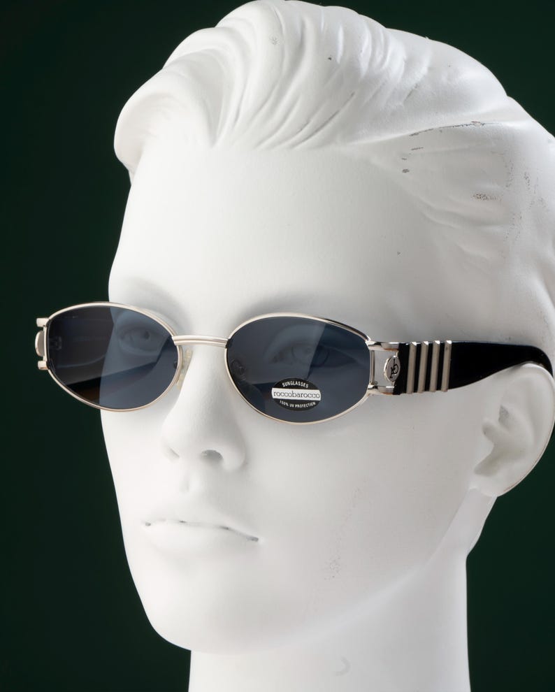 May include: Oval-shaped sunglasses with silver frames and dark lenses. The temples are black with silver stripes. The sunglasses are displayed on a white mannequin head against a dark green background.