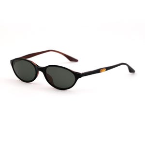 May include: Oval-shaped sunglasses with a black frame and dark lenses. The temples are black with a gold accent near the hinge, and the arms are a dark brown color. The sunglasses are set against a white background.