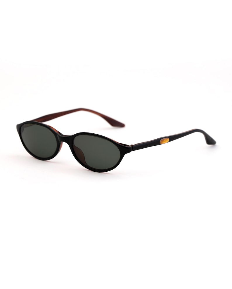 May include: Oval-shaped sunglasses with a black frame and dark lenses. The temples are black with a gold accent near the hinge, and the arms are a dark brown color. The sunglasses are set against a white background.