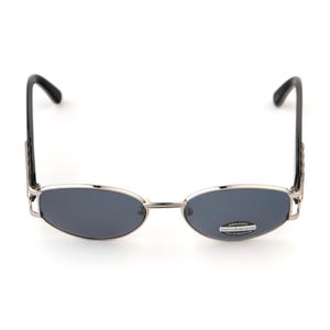 May include: Oval-shaped sunglasses with a silver frame and dark gray lenses. The temples are black and feature a decorative design. The sunglasses have a small sticker on the lens.
