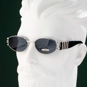 May include: Oval-shaped sunglasses with dark lenses and a silver frame. The temples are black with silver stripes. The sunglasses are displayed on a white bust against a dark green background. The lenses have a small sticker with text.