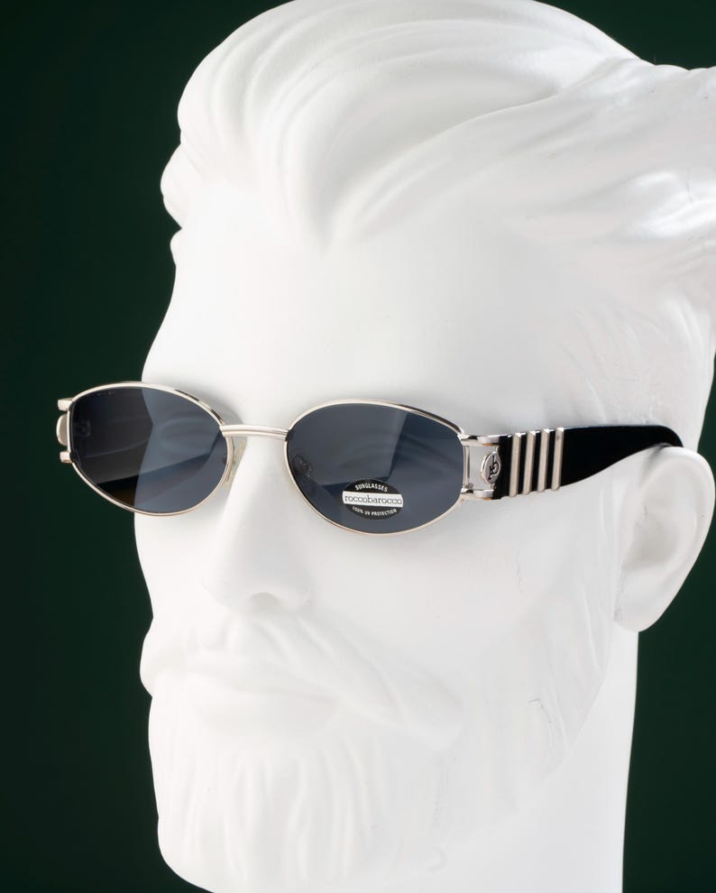 May include: Oval-shaped sunglasses with dark lenses and a silver frame. The temples are black with silver stripes. The sunglasses are displayed on a white bust against a dark green background. The lenses have a small sticker with text.