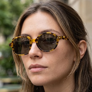 Pode incluir: Óculos de sol com armação tartaruga e lentes escuras. A armação apresenta um design misto oval e quadrado, com tons de castanho e amarelo. Usados por uma pessoa com cabelo castanho claro.