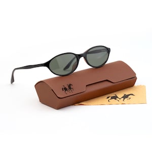 May include: Black oval sunglasses with dark green lenses rest on a brown leather case. A tan cleaning cloth with a horse logo is next to the case. The sunglasses have a classic design, suitable for everyday wear.