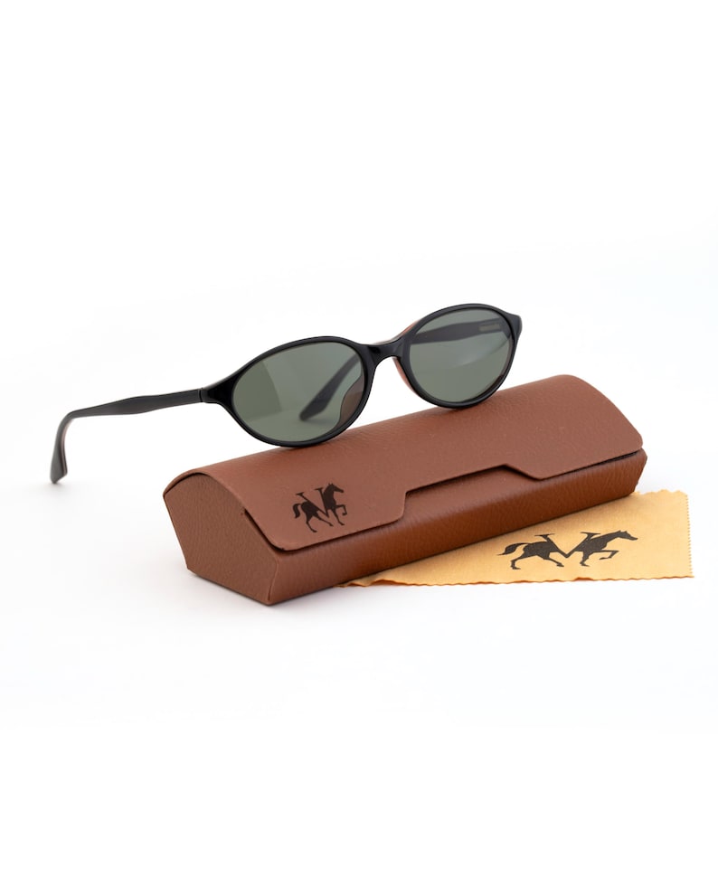 May include: Black oval sunglasses with dark green lenses rest on a brown leather case. A tan cleaning cloth with a horse logo is next to the case. The sunglasses have a classic design, suitable for everyday wear.