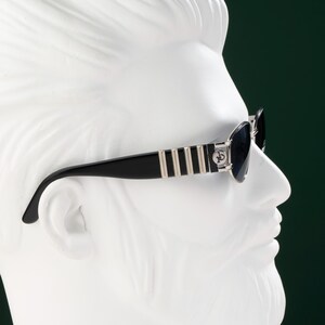 May include: Black sunglasses with silver accents and a decorative emblem on the temples. The sunglasses are set against a white bust with a dark green background. The frames have a striped design.
