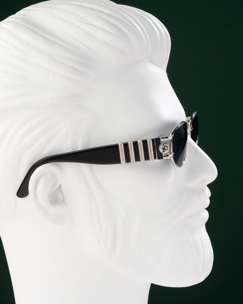 May include: Black sunglasses with silver accents and a decorative emblem on the temples. The sunglasses are set against a white bust with a dark green background. The frames have a striped design.