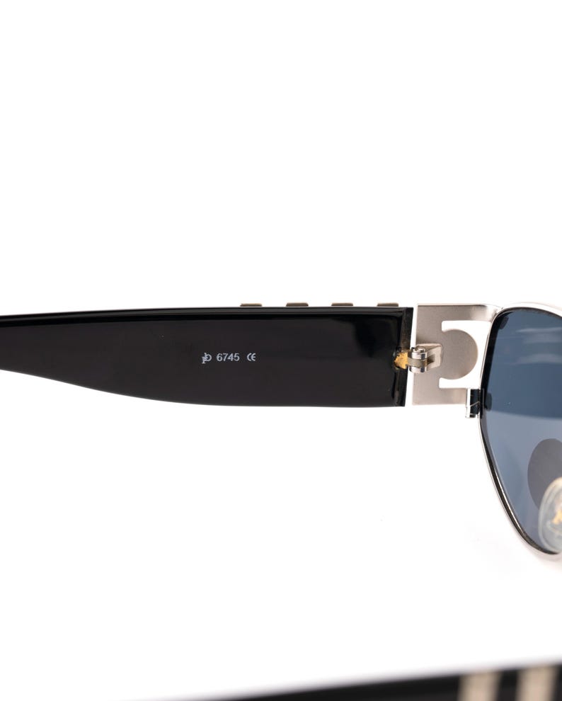 May include: Black sunglasses with a silver metal accent on the temple. The sunglasses have dark blue lenses and the text "6745 CE" is printed on the black arm.