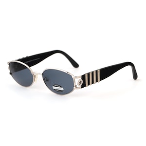 May include: Oval-shaped sunglasses with a silver frame and dark lenses. The temples are black with silver vertical stripes. The sunglasses have a classic, retro design.