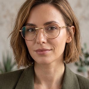 May include: A woman wearing round eyeglasses with a gold frame and a tortoiseshell pattern. The woman is wearing a green blazer. The background is a neutral color.