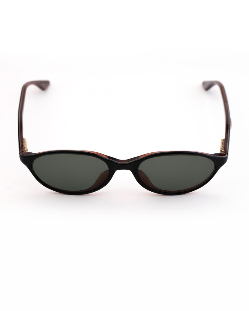 May include: Black oval sunglasses with dark lenses. The frames are black with brown accents and temples. The sunglasses are set against a white background.