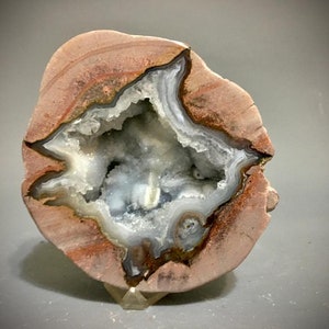 Puede incluir: Una sección transversal de una geoda, que muestra una cavidad llena de cristales brillantes, azul claro y blancos. La cáscara exterior es de color marrón rojizo con patrones en capas. La geoda se exhibe sobre un soporte transparente.