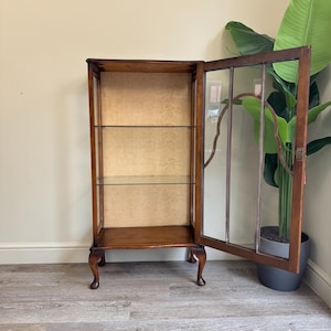 May include: A vintage wooden display cabinet with glass shelves and a patterned back. The cabinet has a dark brown finish, curved legs, and a glass door ajar. The cabinet's dimensions are approximately 150cm in height. A large green plant is in the background.