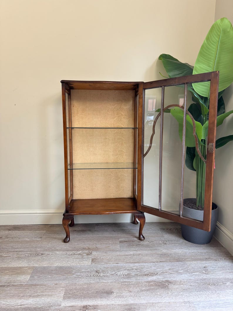 May include: A vintage wooden display cabinet with glass shelves and a patterned back. The cabinet has a dark brown finish, curved legs, and a glass door ajar. The cabinet's dimensions are approximately 150cm in height. A large green plant is in the background.