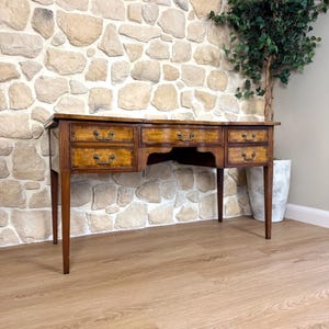 May include: A wooden desk featuring a rectangular top and five drawers with ornate metal pulls. The desk has tapered legs and a warm brown finish with lighter wood grain detailing on the drawer fronts. The structure is symmetrical with two drawers on each side and one central drawer.