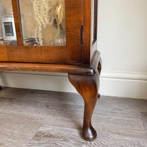 May include: A dark wood display cabinet with glass panels, displaying dried flowers and a candle. The cabinet features a curved leg design and a rich, polished finish. The interior is visible through the glass, offering a glimpse of the contents.