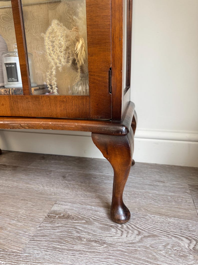 May include: A dark wood display cabinet with glass panels, displaying dried flowers and a candle. The cabinet features a curved leg design and a rich, polished finish. The interior is visible through the glass, offering a glimpse of the contents.