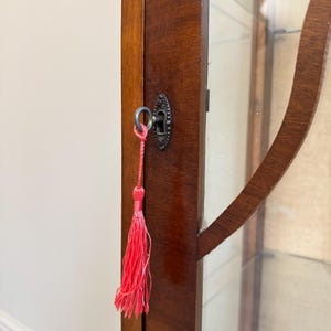 May include: A detail shot of a vintage wooden display cabinet with a glass door. The cabinet is finished in a dark brown stain and features a decorative keyhole with a bright pink tassel. The cabinet is likely for displaying collectibles.