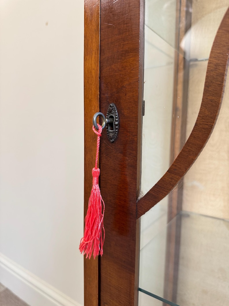 May include: A detail shot of a vintage wooden display cabinet with a glass door. The cabinet is finished in a dark brown stain and features a decorative keyhole with a bright pink tassel. The cabinet is likely for displaying collectibles.