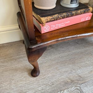 May include: A dark brown wooden side table with a curved leg. The table has a shelf with a stack of books, a beige pot, and a candle. The book on top is black, and the book below is pink with the title "The Monocle Guide to Cosy Homes."