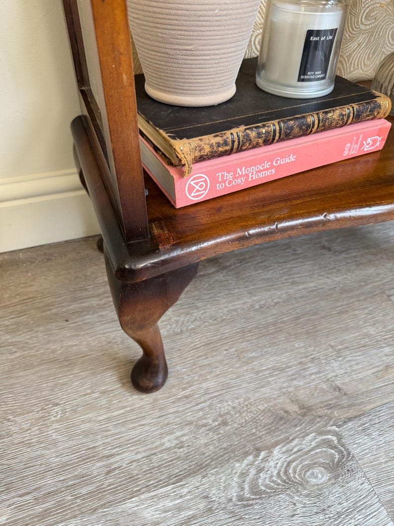 May include: A dark brown wooden side table with a curved leg. The table has a shelf with a stack of books, a beige pot, and a candle. The book on top is black, and the book below is pink with the title "The Monocle Guide to Cosy Homes."