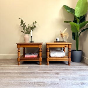 May include: Two wooden side tables with a lower shelf and turned legs. One table has a vase with greenery and a candle, the other has a white blanket. A large potted plant is in the background.