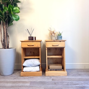 May include: Two wooden bedside tables with drawers and shelves. The bedside tables are made of light wood and have a simple design. One bedside table has a white pillow and blanket on the shelf, while the other has a book and a box on the shelf.