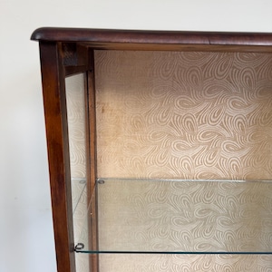 May include: A dark wood display cabinet with glass shelves and a patterned backing. The cabinet has a curved top. The interior features a light beige, swirling pattern. The glass shelves are clear and reflect light. The cabinet is likely for displaying collectibles.