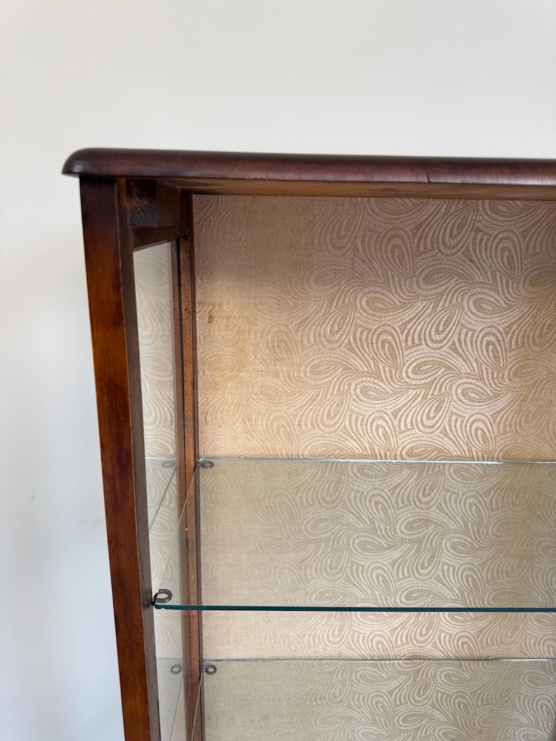 May include: A dark wood display cabinet with glass shelves and a patterned backing. The cabinet has a curved top. The interior features a light beige, swirling pattern. The glass shelves are clear and reflect light. The cabinet is likely for displaying collectibles.