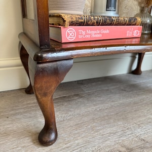 May include: A dark wooden side table with a glass door, displaying two books and a woven basket on its shelf. The table has curved legs and a rich brown finish. A candle and vase are visible on the top.
