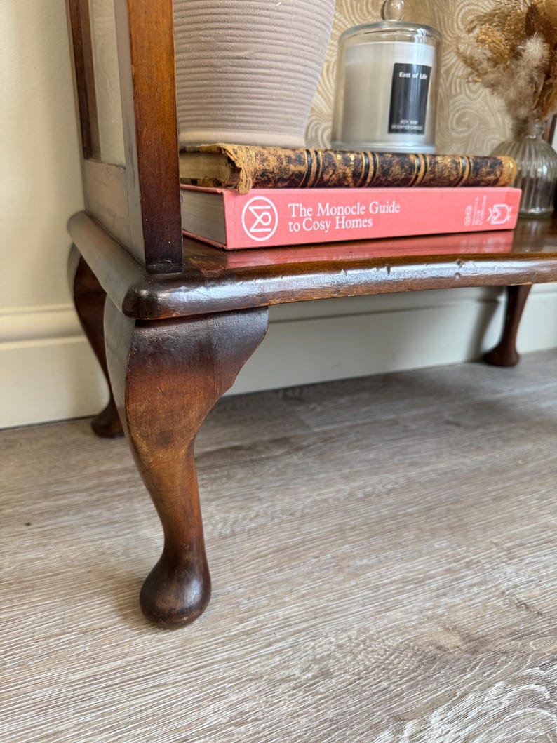 May include: A dark wooden side table with a glass door, displaying two books and a woven basket on its shelf. The table has curved legs and a rich brown finish. A candle and vase are visible on the top.