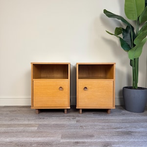 May include: A pair of light oak bedside cabinets. Each cabinet has a single drawer with a round handle and an open shelf. The cabinets are on short legs and have a simple, minimalist design.
