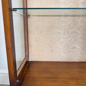 May include: A wooden display cabinet with glass shelves and a patterned interior. The cabinet has a dark brown finish and a curved base. The interior features a light beige wallpaper with a swirling design.