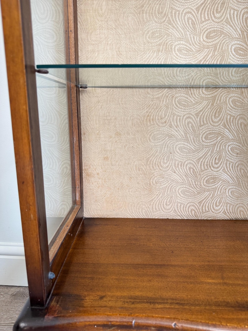May include: A wooden display cabinet with glass shelves and a patterned interior. The cabinet has a dark brown finish and a curved base. The interior features a light beige wallpaper with a swirling design.