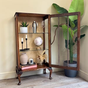 May include: A dark wood display cabinet with glass shelves, partially open to reveal interior contents. The cabinet houses a variety of decorative items: a plant, cocktail shaker, globe, candles, and a vase. The cabinet is set against a patterned backdrop.