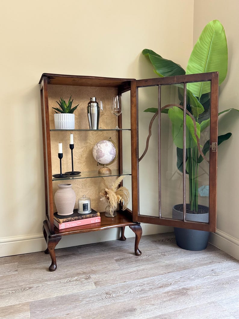 May include: A dark wood display cabinet with glass shelves, partially open to reveal interior contents. The cabinet houses a variety of decorative items: a plant, cocktail shaker, globe, candles, and a vase. The cabinet is set against a patterned backdrop.