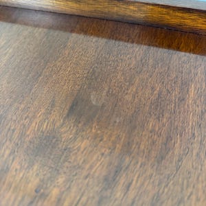 May include: Close-up of a wooden surface with a rich, dark brown colour and visible wood grain. The surface has a smooth, polished appearance, with a darker brown border at the top. The wood appears to be well-maintained.