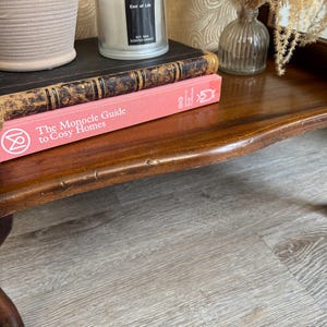 May include: A polished dark wood table displaying a stack of books, a candle in a glass jar, and a small vase with dried flowers. The top book is black with gold detailing, and the bottom book is pink, titled "The Monocle Guide to Cosy Homes".