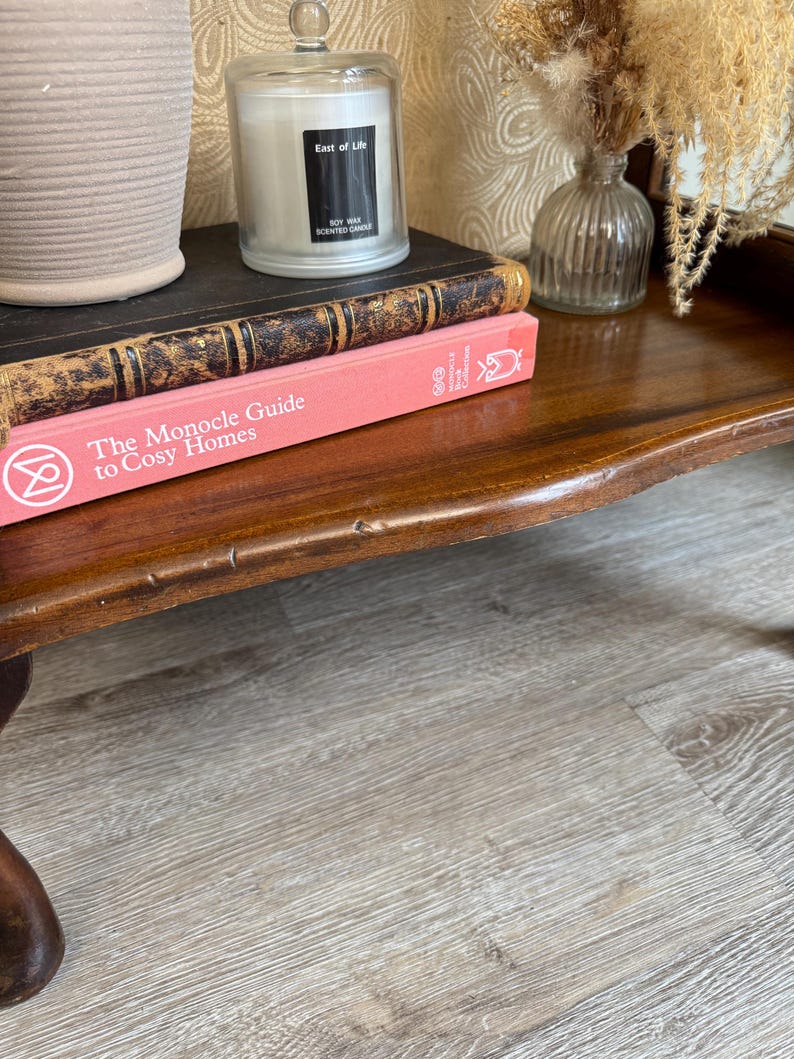 May include: A polished dark wood table displaying a stack of books, a candle in a glass jar, and a small vase with dried flowers. The top book is black with gold detailing, and the bottom book is pink, titled "The Monocle Guide to Cosy Homes".