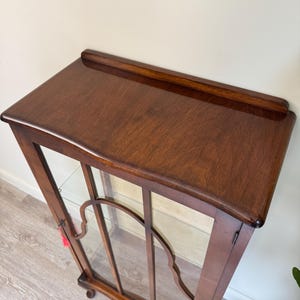 May include: A dark brown wooden display cabinet with a glass front. The cabinet has a curved top and decorative glass panel. The cabinet has a small, rounded foot and a shelf inside. The cabinet is empty. The cabinet is made of dark brown wood.