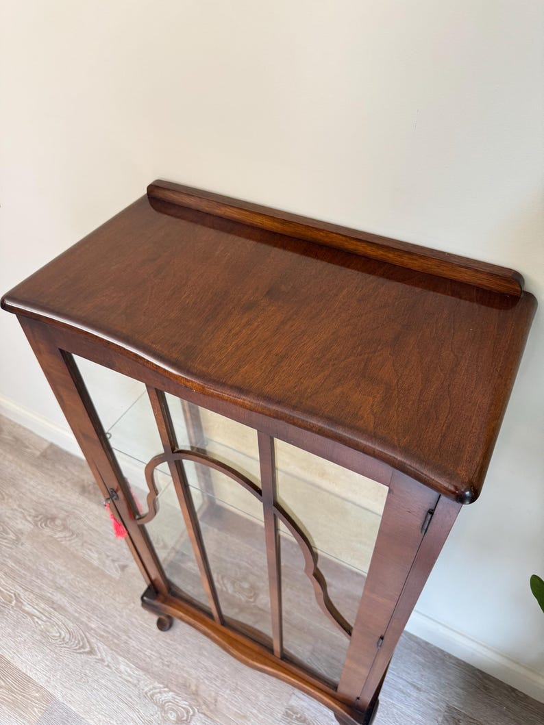 May include: A dark brown wooden display cabinet with a glass front. The cabinet has a curved top and decorative glass panel. The cabinet has a small, rounded foot and a shelf inside. The cabinet is empty. The cabinet is made of dark brown wood.