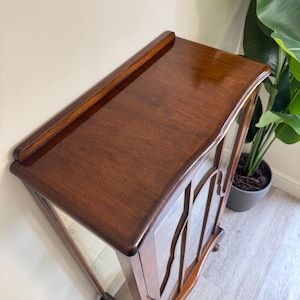 May include: A dark brown wooden display cabinet with glass doors. The cabinet has a glossy finish and decorative wooden detailing on the glass doors. It features a curved top and rests on small feet. The cabinet is likely vintage.