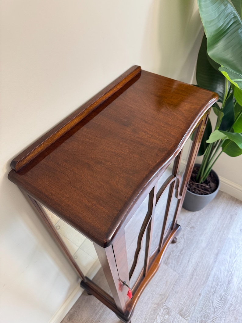 May include: A dark brown wooden display cabinet with glass doors. The cabinet has a glossy finish and decorative wooden detailing on the glass doors. It features a curved top and rests on small feet. The cabinet is likely vintage.