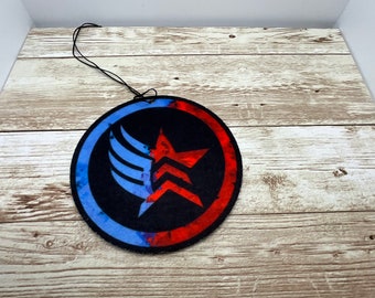 May include: A round, black air freshener with a design featuring a red star and blue wing-like shapes. The design is set within a red and blue circle. A black string is attached for hanging.